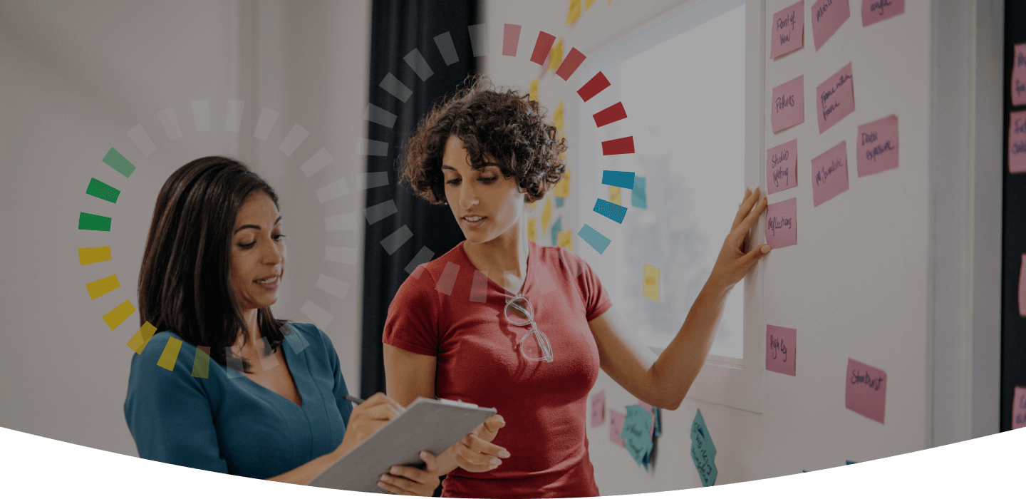 Close-up of two women working together at a whiteboard with sticky notes. Both have DiSC style halos above their heads.