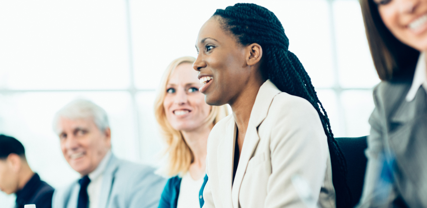 Group of smiling people wearing business attire sitting in a meeting room with the focus on one smiling woman wearing a cream blazer.