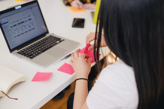 Women taking notes on pink sticky notes while working on laptop.