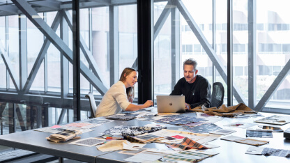 Two coworkers working at a large table with fabric samples. a laptop, and paper spread out all around them.